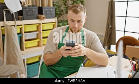 Jeune homme barbu dans le tablier vert concentré sur smartphone à l'intérieur de studio d'espace de travail d'artisanat. Banque D'Images