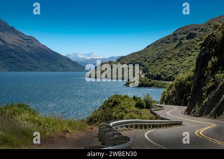 Lac Wakatipu et route sinueuse vide au bord du lac - ensemble contre un ciel bleu clair et des montagnes couvertes d'arbres verts Banque D'Images