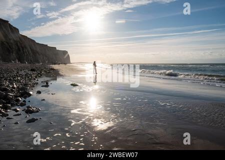 La photographie capture l'essence d'un voyage solitaire le long d'une côte de galets. Un individu solitaire, pris dans l'acte de marcher, se dessine contre les reflets étincelants du soleil sur la surface de l'eau. Le soleil, haut au-dessus, jette un chemin doux et brillant à travers la mer, invitant le spectateur à réfléchir sur le chemin du vagabond. Les falaises offrent une toile de fond majestueuse à cette scène de solitude contemplative. Réflexions sur un voyage côtier. Photo de haute qualité Banque D'Images