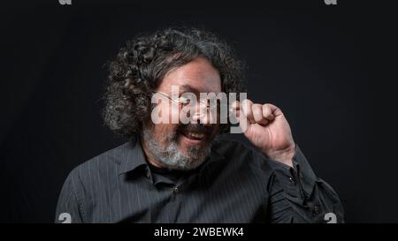 Portrait d'homme latin à la barbe blanche et aux cheveux bouclés noirs avec une expression flirtative, tenant ses lunettes, portant une chemise noire contre un dos noir Banque D'Images
