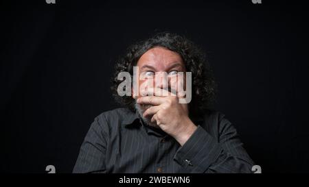 Portrait d'homme latin avec barbe blanche et cheveux bouclés noirs avec une expression drôle, couvrant sa bouche avec la main, portant une chemise noire contre bac noir Banque D'Images