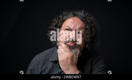 Portrait d'homme latin avec barbe blanche et cheveux bouclés noirs avec une expression indécise, main sur le menton, portant une chemise noire sur fond noir Banque D'Images