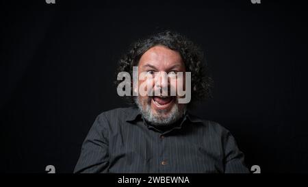 Portrait d'homme latin avec barbe blanche et cheveux bouclés noirs avec expression heureuse et excitée, portant une chemise noire sur fond noir Banque D'Images