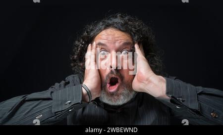 Portrait d'homme latin avec barbe blanche et cheveux bouclés noirs avec une expression surprise, mains sur le visage, portant une chemise noire sur fond noir Banque D'Images