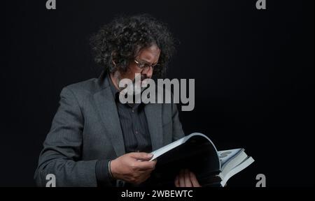 Portrait d'homme latin avec barbe blanche et cheveux bouclés noirs lisant un livre, portant une chemise noire sur fond noir Banque D'Images