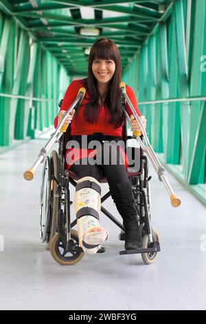 Une jeune femme avec une jambe en plâtre est assise dans un fauteuil roulant et sourit, à l'hôpital Banque D'Images