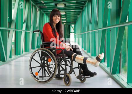 Une jeune femme avec une jambe en plâtre est assise dans un fauteuil roulant et sourit, à l'hôpital Banque D'Images