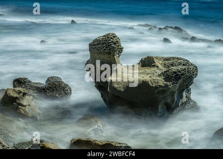 Une photo longue exposition capture le mouvement serein des vagues brumeuses tourbillonnant autour de rochers sculptés à motifs en nid d'abeille au bord de la mer Banque D'Images