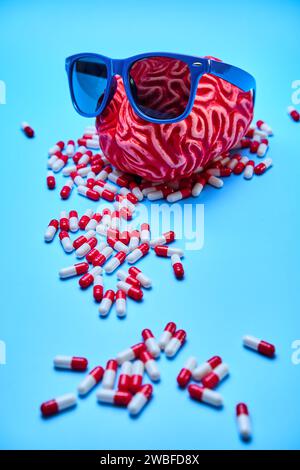 Cerveau rouge avec des lunettes de soleil sur un tas de capsules rouges et blanches sur une surface bleue. Banque D'Images
