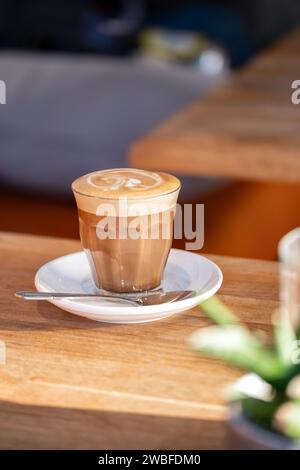 Un café cappuccino fraîchement préparé, servi dans un verre sur une soucoupe dans un café. Le cappuccino a une garniture pleine de mousse de lait décorée avec de l'art de latte Banque D'Images