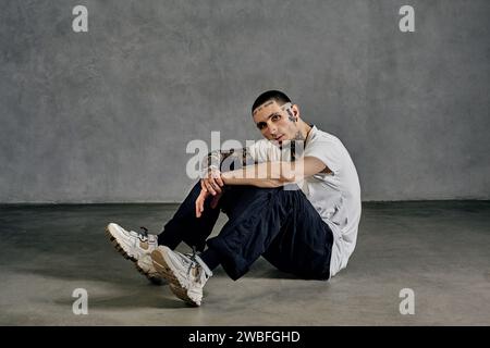 Tatoué avec barbe. Habillé en t-shirt blanc, baskets, pantalon de sport noir. Assis latéralement sur le sol, fond gris. Dancehall, hip-hop Banque D'Images