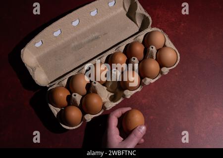 Douzaine d'œufs bruns de la boîte en carton et main tenant un œuf sur un fond rouge vif avec une texture intéressante Banque D'Images