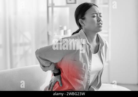 Triste femme asiatique assise sur le canapé, touchant le dos, photo monochrome Banque D'Images