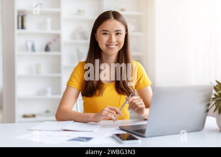 Jeune femme asiatique assise au bureau avec ordinateur portable à la maison et souriant Banque D'Images