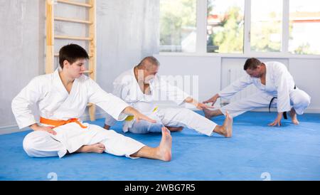 Hommes d'âges différents dans un kimono blanc faisant des étirements musculaires avant l'entraînement de karaté ou de judo Banque D'Images