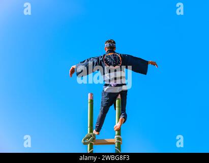 Un pompier hikeshi japonais debout sur une jambe au sommet d'une grande échelle de bambou effectuant des acrobaties pendant le spectacle traditionnel Hashigo-nori o Banque D'Images