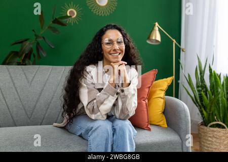 Une femme joyeuse en vêtements décontractés profite d'un moment confortable assise sur un canapé gris, entourée de plantes d'intérieur vibrantes et d'un décor chic. Banque D'Images