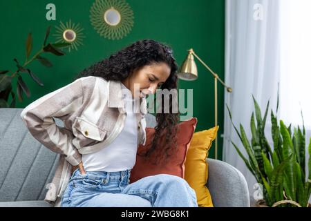 Fatigué surchargé de travaux ménagers et femme de ménage, femme hispanique assise dans le cinéma de salon tenant les mains derrière le dos, mal de dos. Banque D'Images