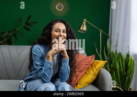 Une femme joyeuse aux cheveux bouclés porte une chemise en denim tout en étant confortablement assise sur un canapé gris, entourée de plantes d'intérieur vibrantes dans un salon chaleureusement décoré. Banque D'Images