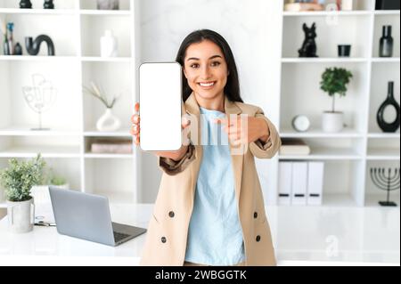 Femme arabe ou indienne brunette positive réussie, pdg d'entreprise, gestionnaire, en costume élégant, stand dans le bureau moderne, montrant le téléphone portable avec écran de maquette blanc pour la publicité ou la présentation, sourire Banque D'Images