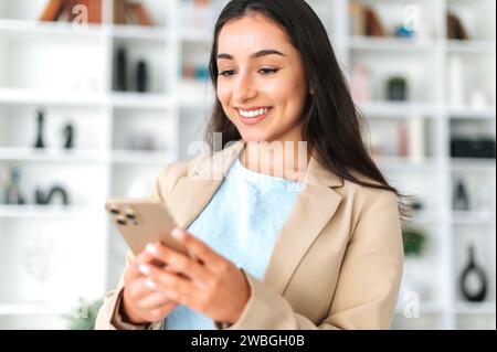 La femme utilise un téléphone intelligent. Positive femme arabe ou indienne réussie dans un costume, se tient dans le bureau, tenant le téléphone portable, envoyant des SMS dans les médias sociaux avec des amis ou des clients, faisant commande en ligne, sourires Banque D'Images