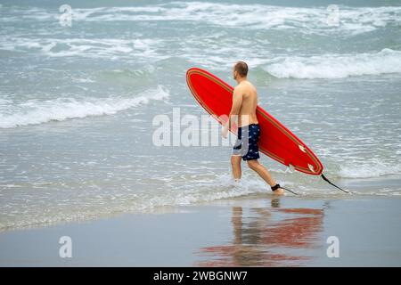 Personne non identifiée entrant dans l'eau, avec une planche de surf rouge. Banque D'Images