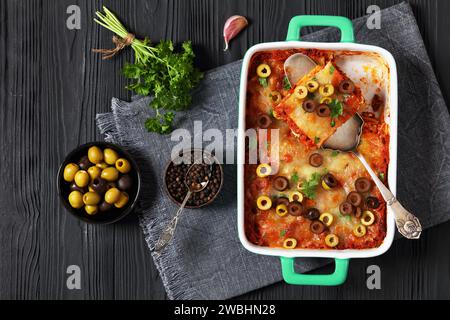 lasagnes garnies de rondelles d'olive et de persil frais dans un plat de cuisson vert sur une table en bois noir avec persil, pelle à gâteau et grains de poivre, horizontal Banque D'Images