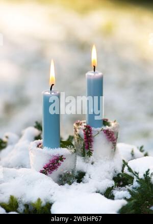 Beaux bougeoirs de Noël en glace, branches roses de fleur de calluna avec des bougies bleues brûlantes dans le garen couvert de neige. Main extérieure d'hiver Banque D'Images