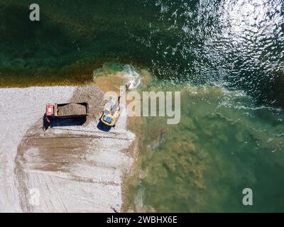 Excavatrice creusant et chargeant du sable dans un camion à benne basculante au bord de la rivière. Machinerie lourde travaillant dans une carrière de sable. Vue aérienne drone de la pelle et du camion. Banque D'Images