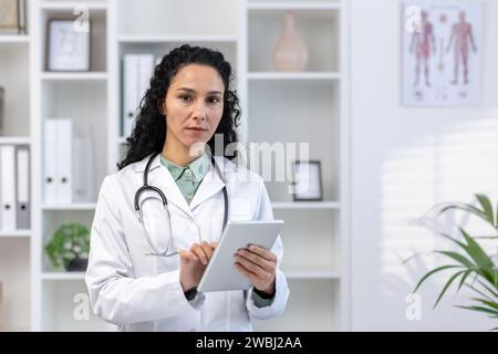 Gros plan portrait de jeune femme médecin, femme latino-américaine utilisant un ordinateur tablette, l'air pensif sérieux et concentré à la caméra, debout à l'intérieur du bureau dans le cabinet médical de la clinique. Banque D'Images