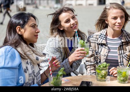 Cette image représente trois femmes plongées dans une conversation animée dans un café de la place de la ville par un jour de fraîcheur. Leurs cheveux sont touffus par le vent, ajoutant un élément dynamique et naturel à la scène. Chaque femme tient un mojito rafraîchissant, contribuant ainsi à un cadre social détendu. La place est animée par l'activité en arrière-plan, mais les femmes sont concentrées sur leur échange, mettant en valeur leur camaraderie intime dans un espace public. Journée venteuse de rattrapage au City Square Cafe. Photo de haute qualité Banque D'Images