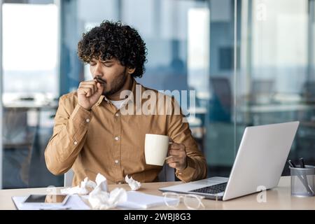 Homme d'affaires malade au travail, homme toussant et éternuant, nez froid et qui coule, assis au bureau à l'intérieur du bureau, tenant une tasse de boisson chaude Banque D'Images