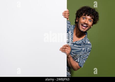 Photo de jeune homme mexicain choqué aux cheveux ondulés se cachant derrière la bannière blanche espace vide pour votre publicité isolé sur fond de couleur kaki Banque D'Images