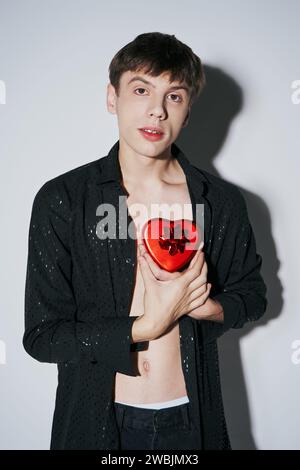 Jeune homme romantique dans la chemise tenant la boîte de cadeau en forme de coeur rouge sur fond gris, Saint Valentin Banque D'Images