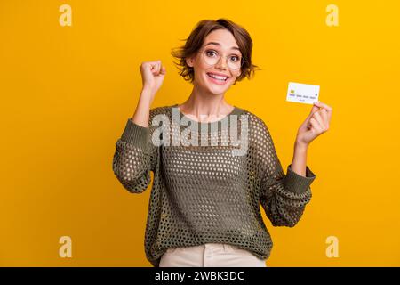 Portrait photo de joli poing de jeune fille célébrer la carte de crédit habillé élégant costume kaki tricoté isolé sur fond de couleur jaune Banque D'Images