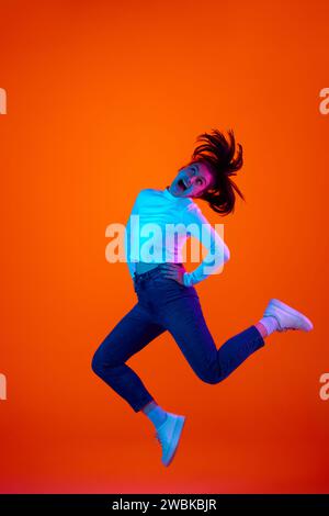 Portrait pleine longueur de jeune femme surjoyée sauter haut de joie et de bonheur sur fond rose-orange dégradé dans la lumière de néon. Banque D'Images