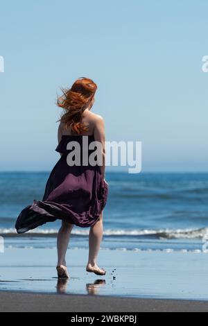 Vue arrière de la femme en robe longue violet foncé, se promenant pieds nus sur la plage de sable fin vers l'eau cristalline de la mer bleue sur la journée ensoleillée avec le ciel clair Banque D'Images