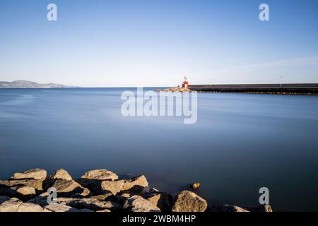 Phare rouge lointain, entouré de rochers et d'une mer bleue calme. Banque D'Images