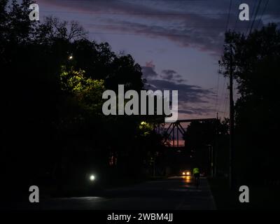 Cycliste monte un vélo le long d'une piste cyclable vers un pont et beau coucher de soleil coloré. Voiture avec phare allumé, arbres éclairés avec des lanternes de rue Banque D'Images