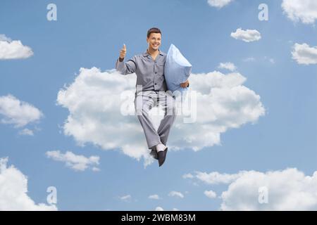Jeune homme en pyjama tenant un oreiller et faisant des gestes pouces vers le haut sur un nuage Banque D'Images