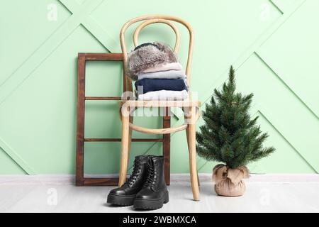 Vêtements empilés sur une chaise avec des bottes et un petit arbre de Noël près du mur en bois vert Banque D'Images