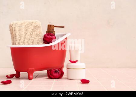 Mini baignoire avec produits cosmétiques, éponge et roses sur tuile rose près du mur blanc. Fête de la Saint-Valentin Banque D'Images