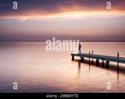 Silhouette d'un homme sur la passerelle d'un lac contemplant un magnifique coucher de soleil. Banque D'Images