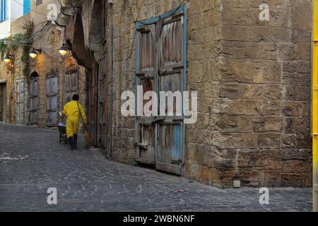 Un nettoyeur d'ordures errant dans une rue vide de Batroun avec de nombreuses portes en bois tôt le matin. Banque D'Images