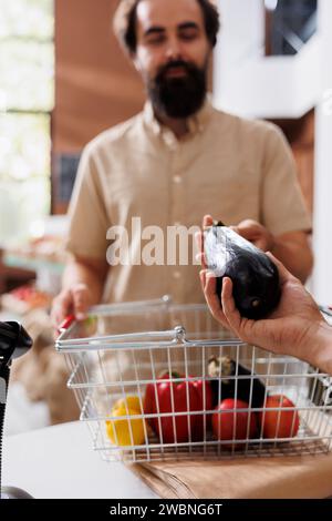 Gros plan d'un homme caucasien barbu donnant de l'aubergine naturelle à un vendeur pour pesée. Client masculin à la caisse pour vérifier le poids des fruits et légumes fraîchement récoltés. Banque D'Images