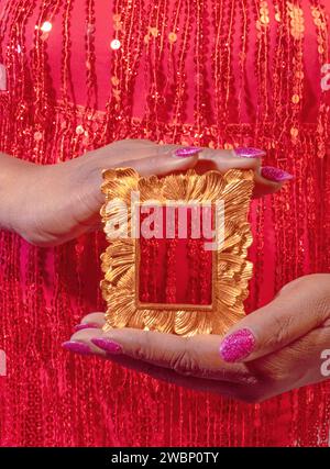 Une femme afro-américaine diversifiée vêtue de sequins rouges et portant un vernis à ongles rose tenant un cadre en or Banque D'Images