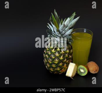 Un grand verre à facettes d'un mélange de jus de fruits sur fond noir, à côté de morceaux d'ananas mûr et de morceaux de kiwi. Gros plan. Banque D'Images