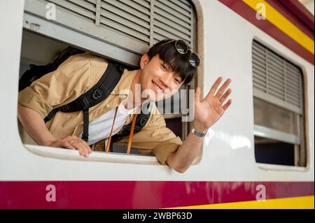 Un jeune voyageur asiatique heureux et excité est dans un train, souriant et agitant sa main, disant au revoir à quelqu'un, et voyageant quelque part par Banque D'Images