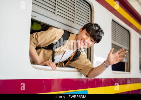 Un jeune voyageur asiatique heureux et excité est dans un train, souriant et agitant sa main, disant au revoir à quelqu'un, et voyageant quelque part par Banque D'Images