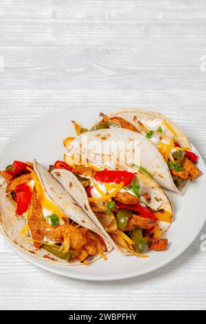 fajitas au poulet tex-mex cuit au four avec poivron mélangé, oignon, crème sure, fromage râpé et tortillas sur une assiette blanche sur une table en bois blanc, verticale Banque D'Images